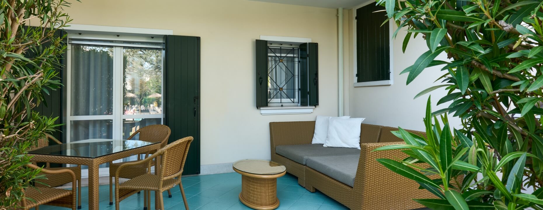 Patio con mobili in rattan, tavolo in vetro e piante verdi.