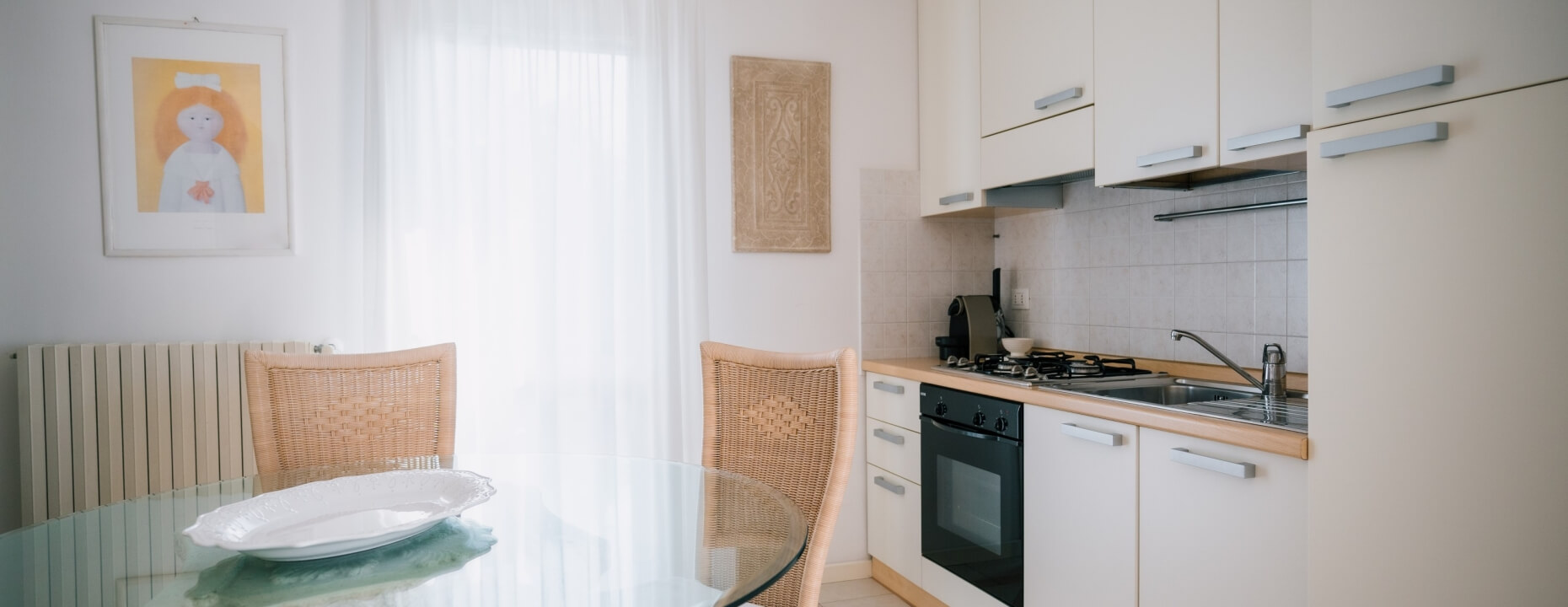 Cucina con armadi bianchi, tavolo in vetro e sedia in vimini.