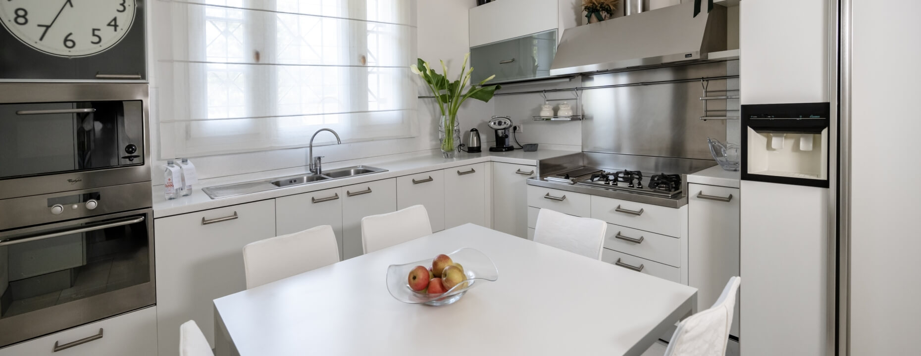 Cucina moderna con mobili bianchi, tavolo, sedie e frutta.