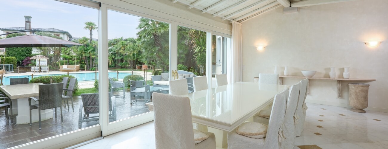 Dining room with a white table, six chairs, and a large window showcasing a pool and garden view.