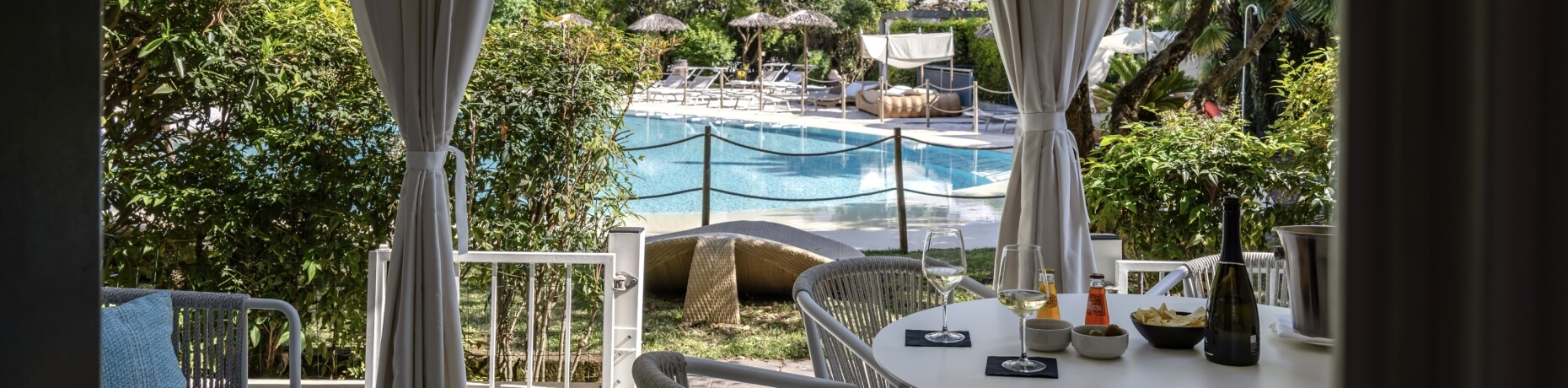 Impostazione di un tavolo per due con vino e snack, piscina blu e verde lussureggiante sullo sfondo.