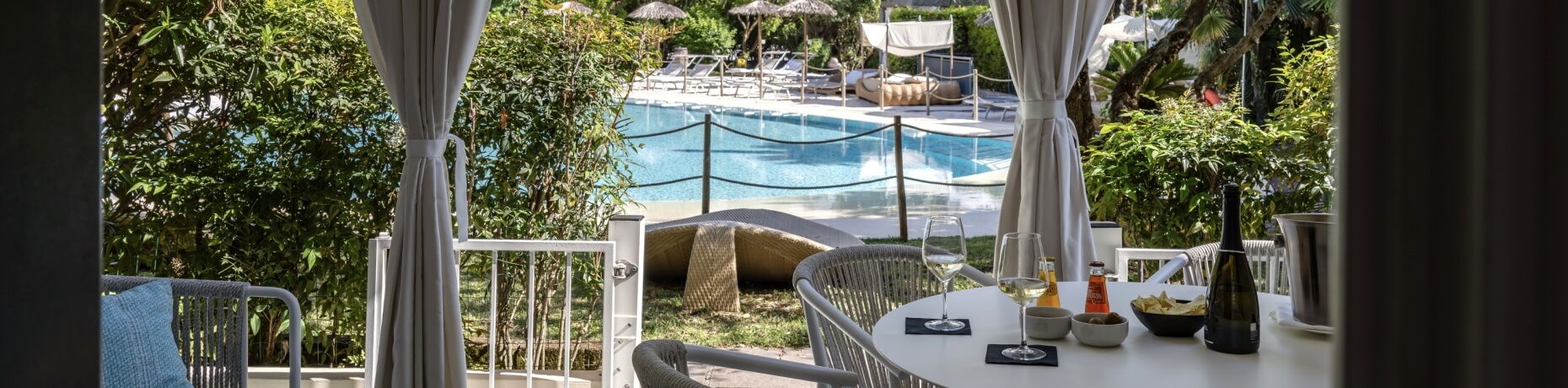 Area esterna soleggiata con tavolo bianco, bicchieri di vino, piscina blu e ombrelloni, circondata da piante verdi.