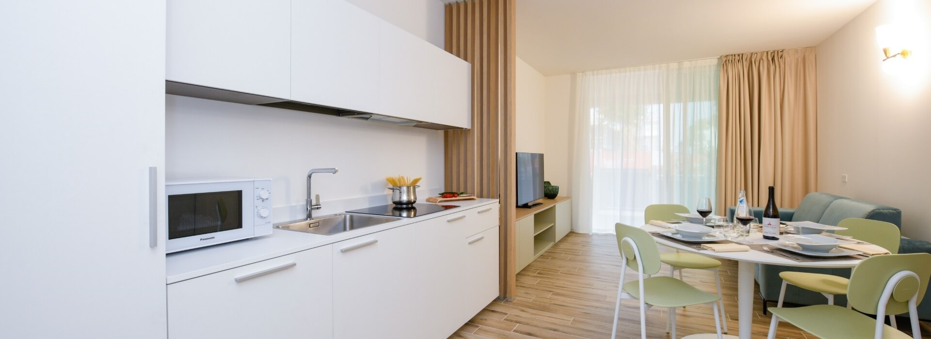 Spazio living moderno con cucina bianca, tavolo da pranzo e sedie verdi, illuminato da luce naturale.