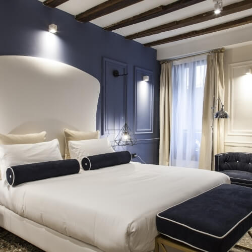 Camera elegante in toni di grigio e bianco, con letto, sgabello tufted e poltrona grigia, illuminata da faretti e lampadario.