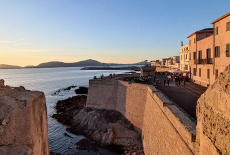 Scena costiera tranquilla con edifici color pesca, persone che passeggiano e un tramonto arancione sullo sfondo.