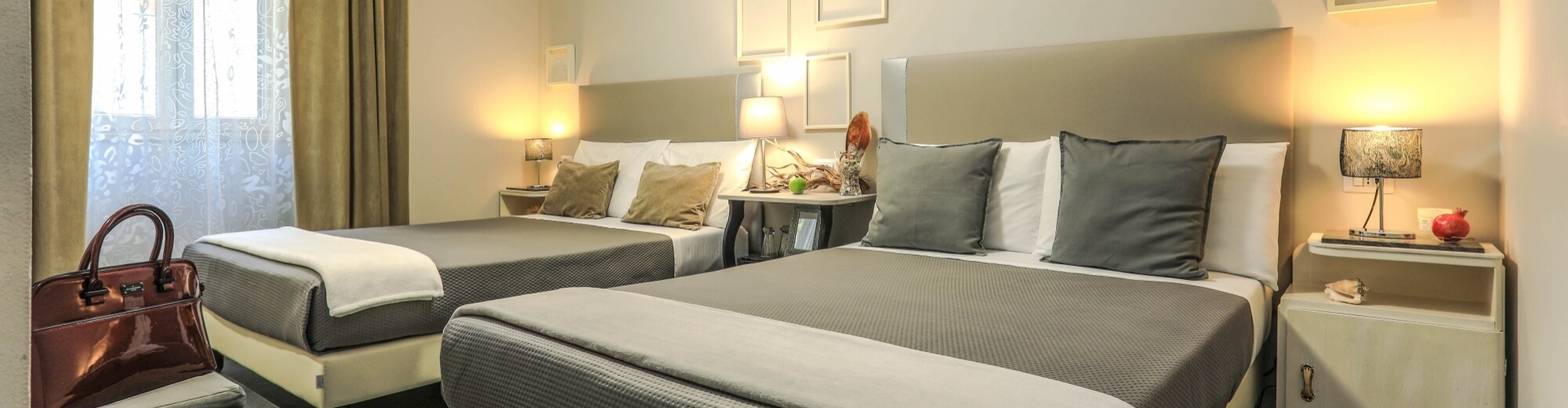 Camera d'hotel con due letti, biancheria bianca, coperte grigie, finestre con tende leggere e decorazioni sulle pareti.
