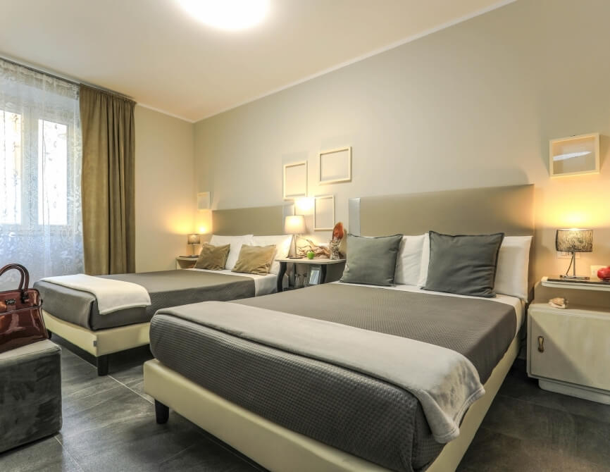 Camera d'albergo con due letti, ottomano, tavolo con lampade, pareti beige e pavimento grigio scuro.