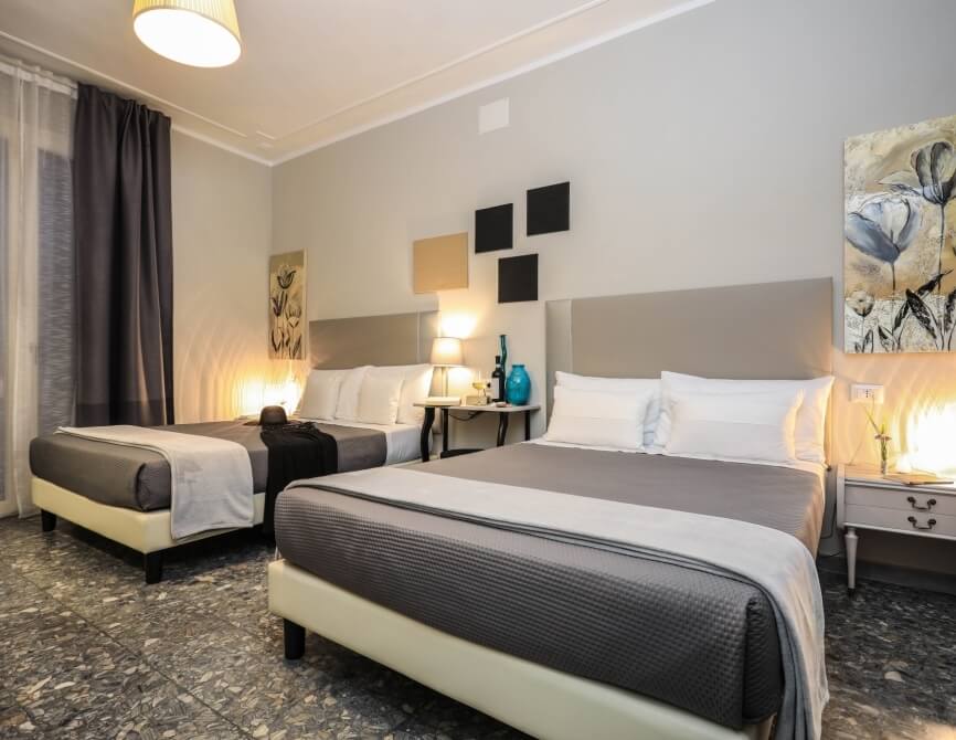 Camera d'hotel con due letti, comodino, panchina, illuminazione soffusa e decorazioni in bianco e grigio.