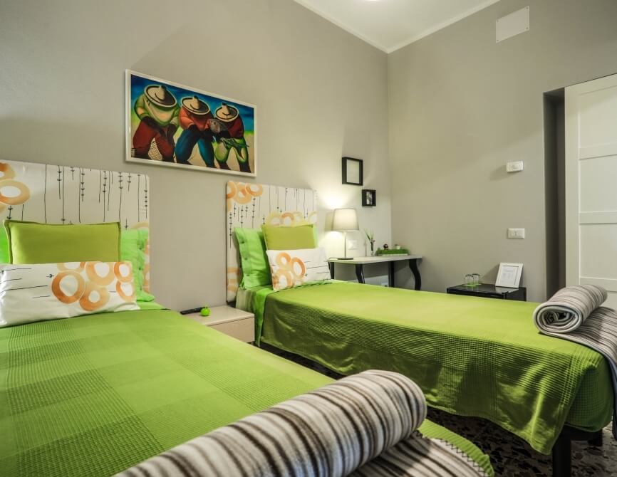 Camera serena con due letti, biancheria verde, lampada, quadro vivace e pareti grigie, trasmette calore e comfort.
