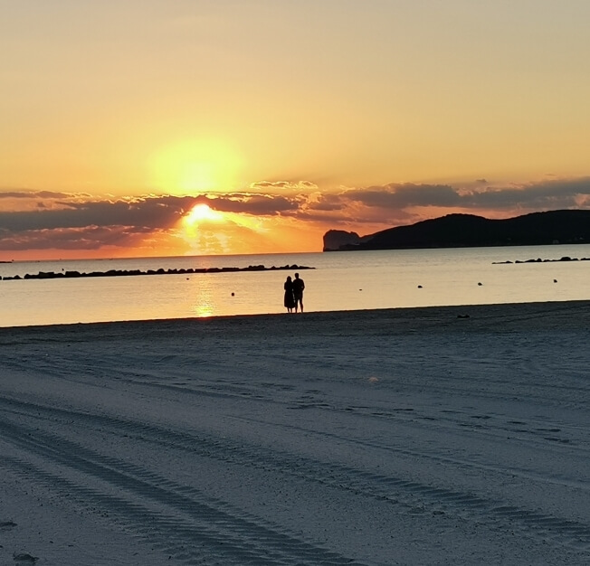 Spiaggia al tramonto con due persone in lontananza, cielo giallo-arancione, jetty roccioso e impronte sulla sabbia.
