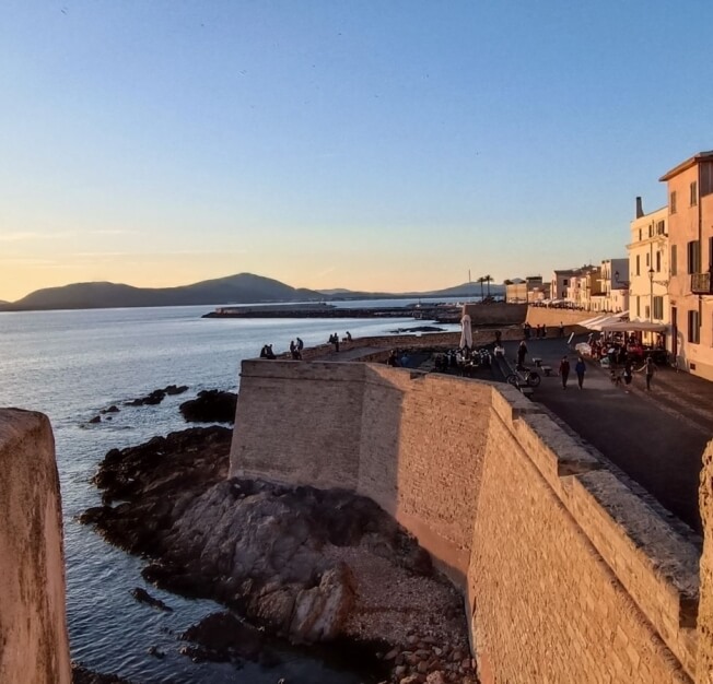Scena costiera serena con un muro di pietra, acqua tranquilla, cielo blu e edifici con persone che socializzano.