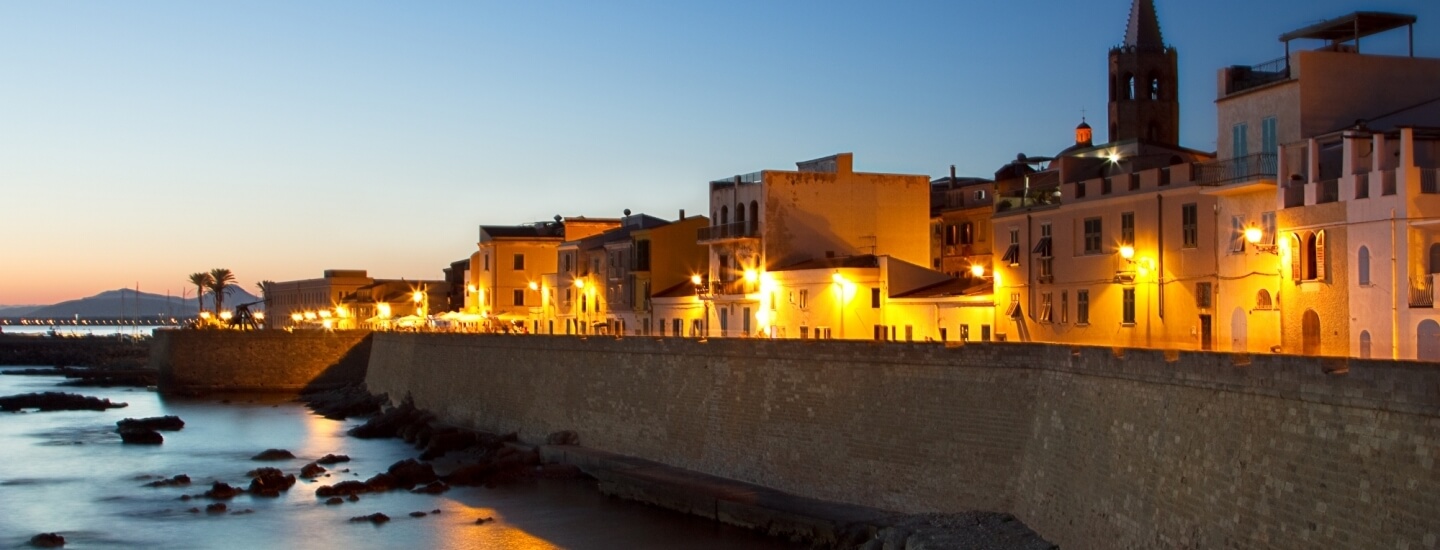 Una tranquilla città costiera al crepuscolo, con un muro di pietra illuminato e edifici gialli e bianchi sullo sfondo montano.