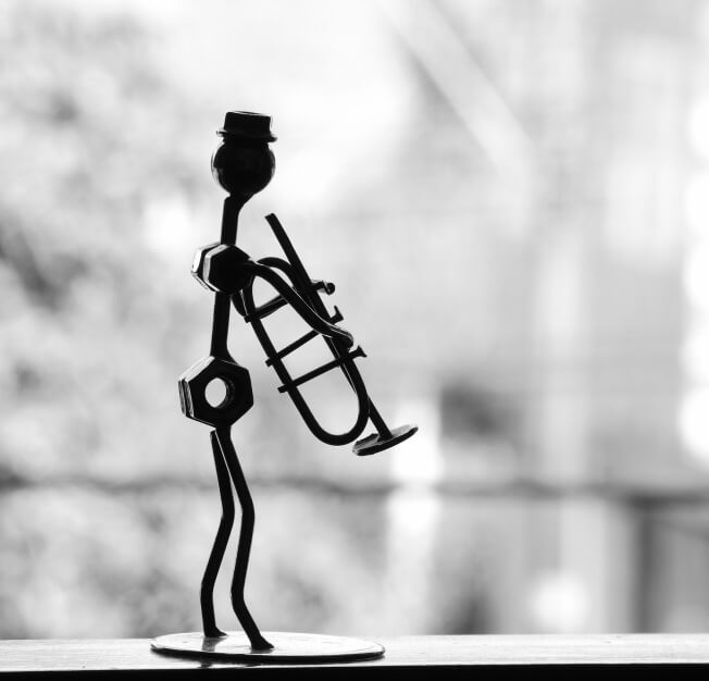 Scultura in metallo di figura slanciata con tromba, su sfondo sfocato di alberi e recinzione, in un'atmosfera serena.