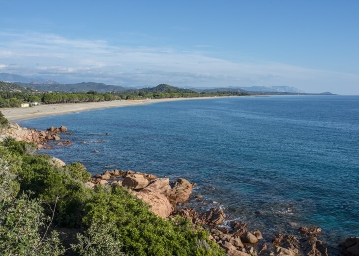 Costa con mare blu, spiaggia sabbiosa e rocce rosse.