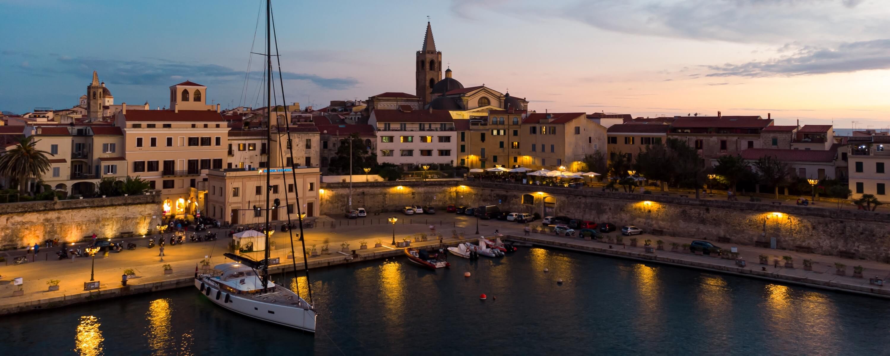 Immagine di un porto al crepuscolo con una barca a vela, luci costiere e un campanile in lontananza.