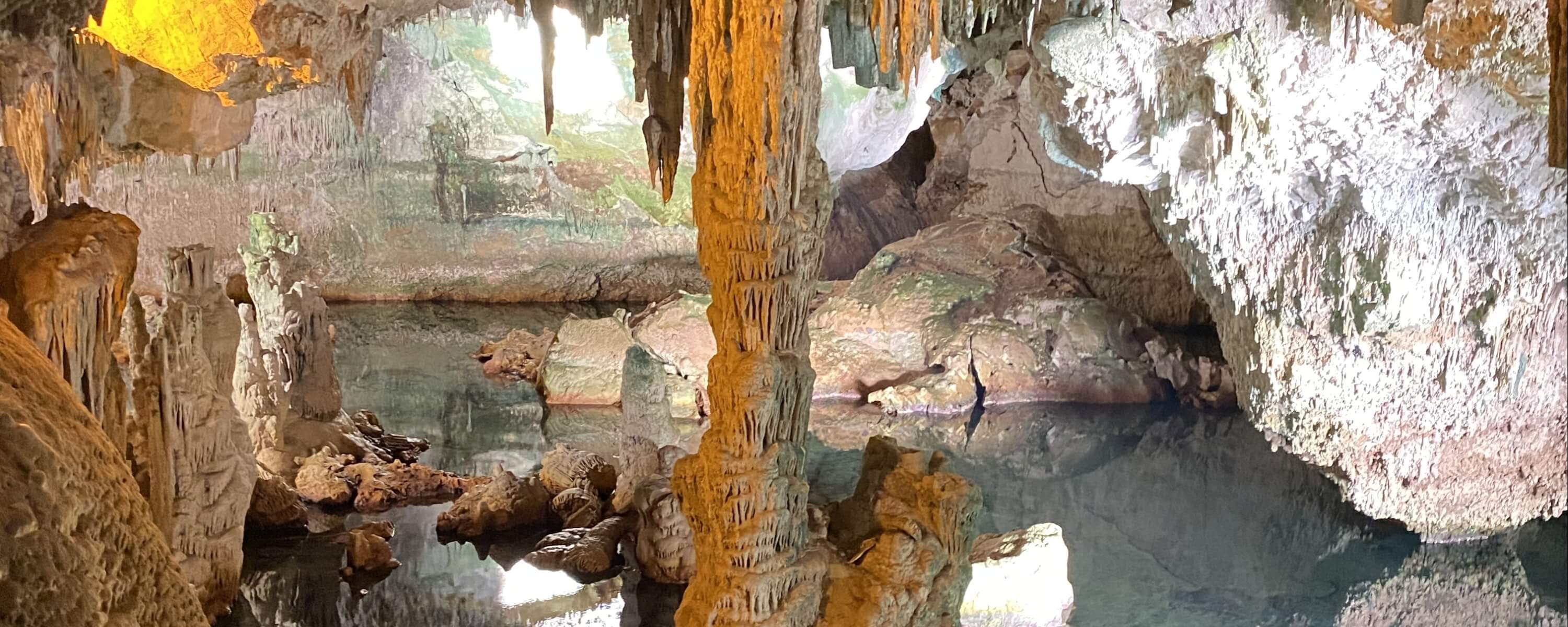 Vista panoramica di una grotta con stalattiti, formazioni rocciose e un lago che riflette il soffitto illuminato.