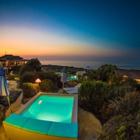 Piscina illuminata con cuscini turchesi, ombrelloni e vista sul tramonto.