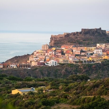 Una vivace città costiera mediterranea con case color pastello, un antico forte e un mare tranquillo sotto cieli nuvolosi.