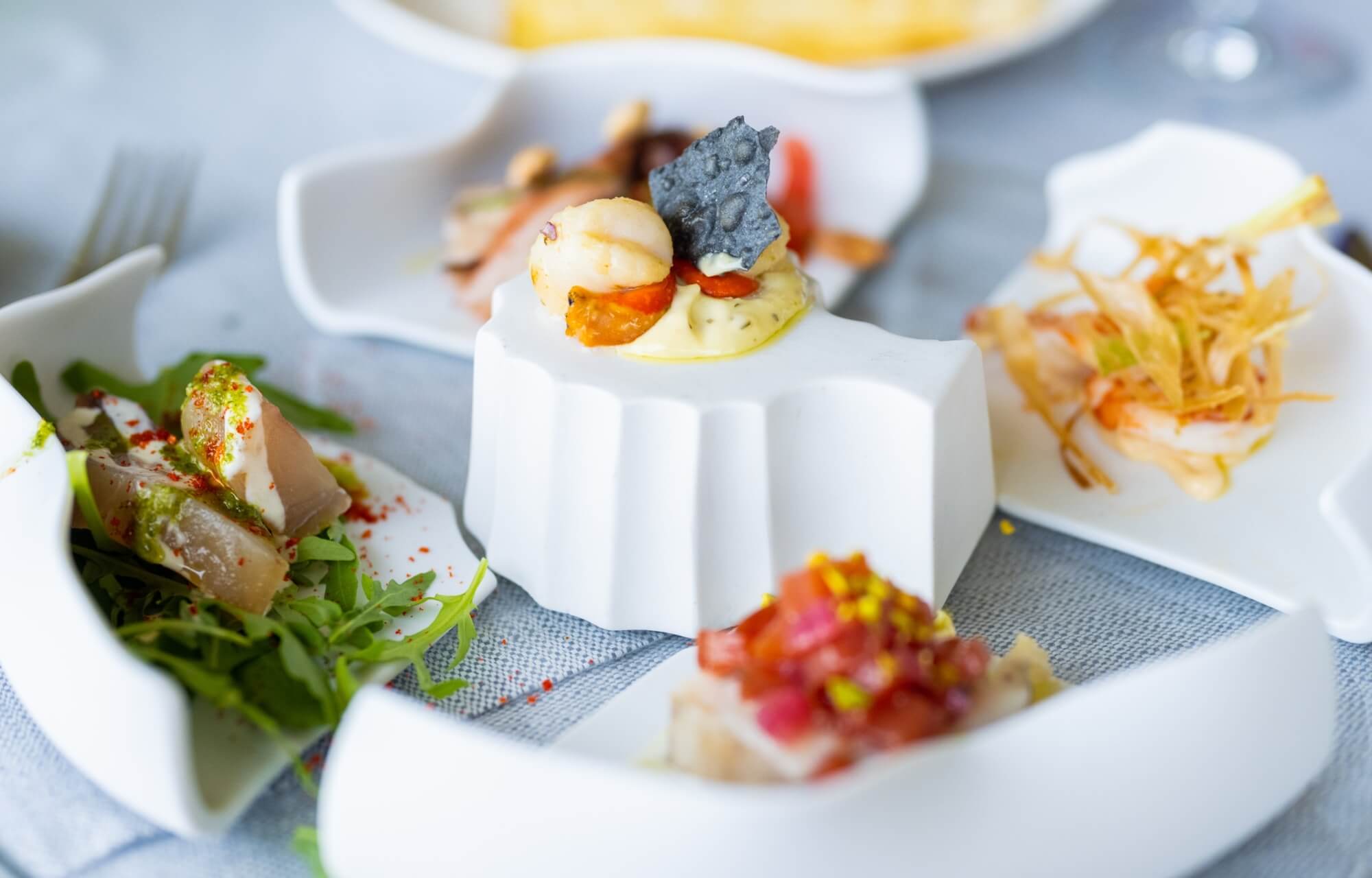 Impostazione della tavola con piatti bianchi irregolari, cibo vario, insalata di rucola e un piatto centrale decorato.