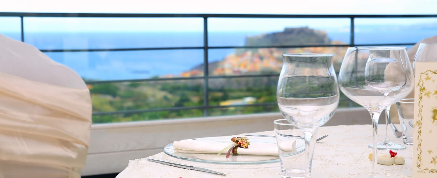 Impostazione della tavola con vista sul mare, tovaglia bianca, piatto in vetro e bicchieri, con una ringhiera nera sullo sfondo.