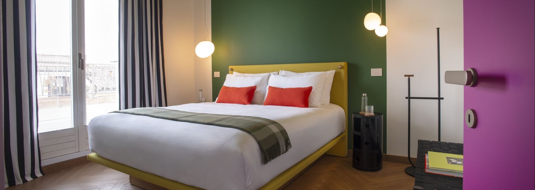 Camera moderna con letto giallo, comodino nero, pareti verdi e bianche, finestra grande e atmosfera rilassante.