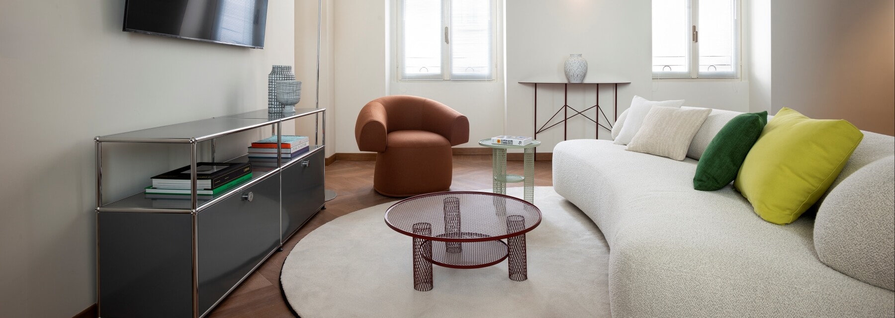 Soggiorno moderno con divano bianco, tavolino rosso, TV, sedia marrone e tavolo console con vaso, illuminato da luce naturale.