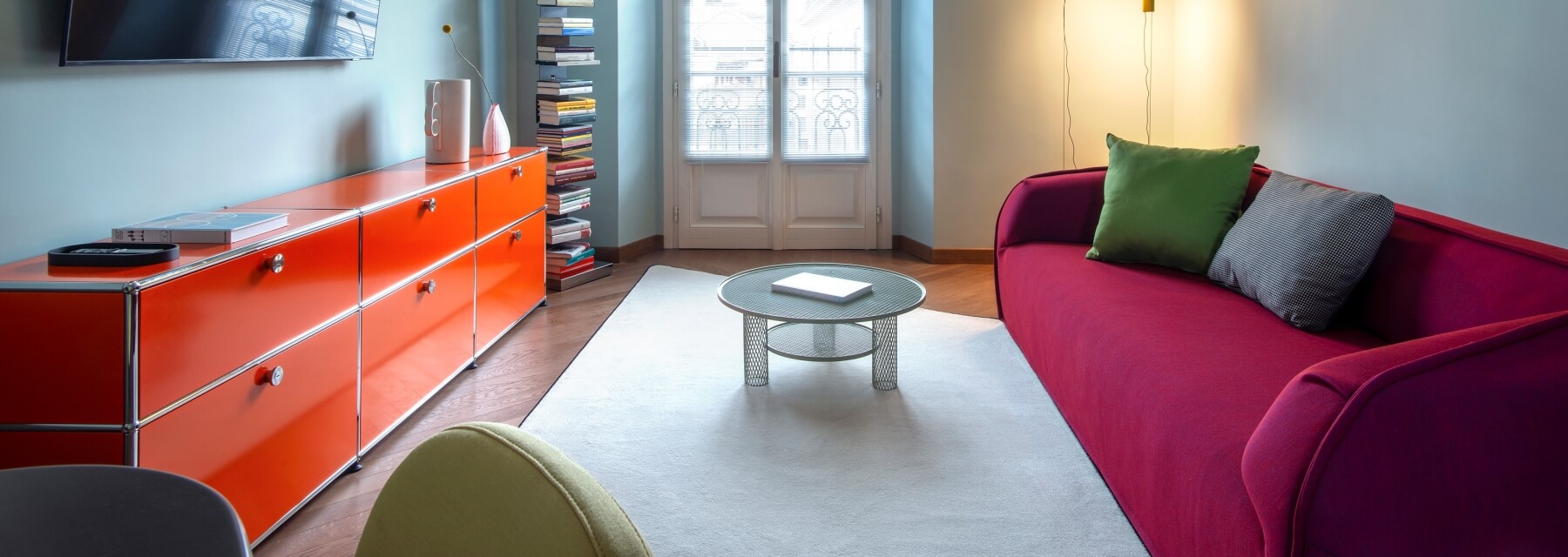 Modern living room with a red couch, white rug, orange dresser, and colorful chairs, illuminated by natural light.