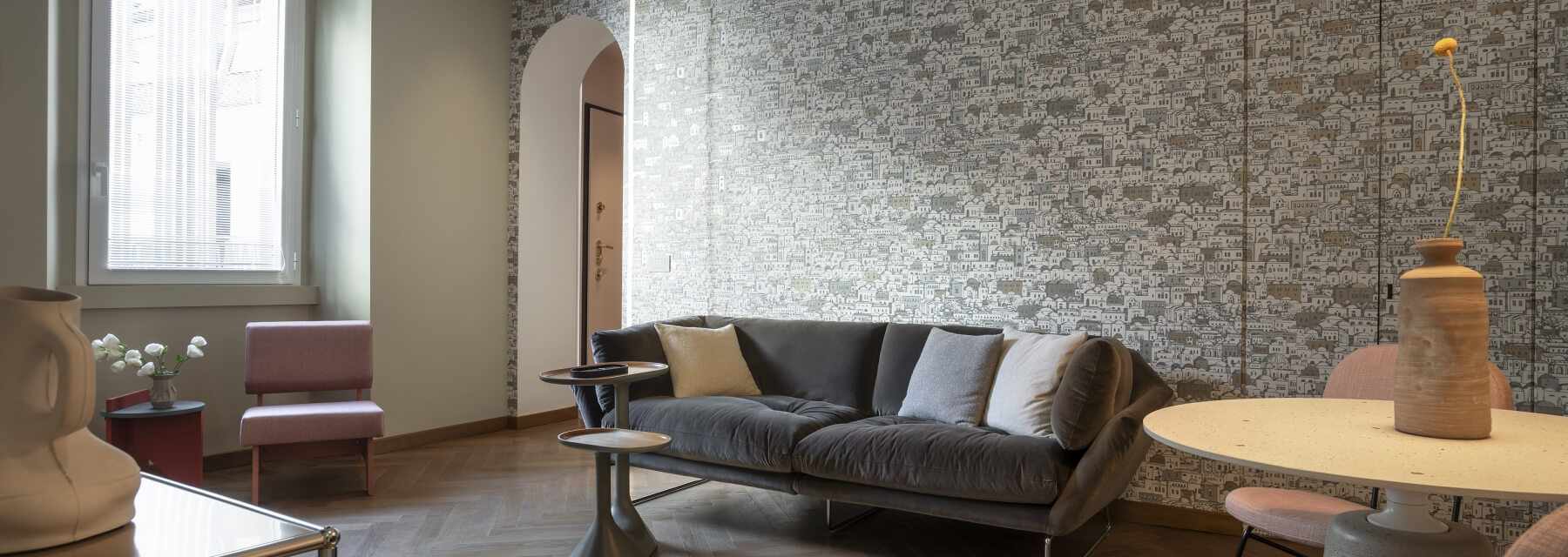 Stylish living room with gray couch, cartoon cityscape wallpaper, asymmetrical coffee table, pink chair, and natural light.
