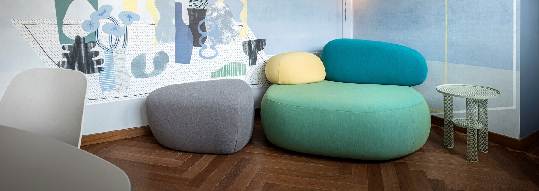 Abstract room with light blue wall mural, dark wood herringbone floor, colorful stools, and a small round table.