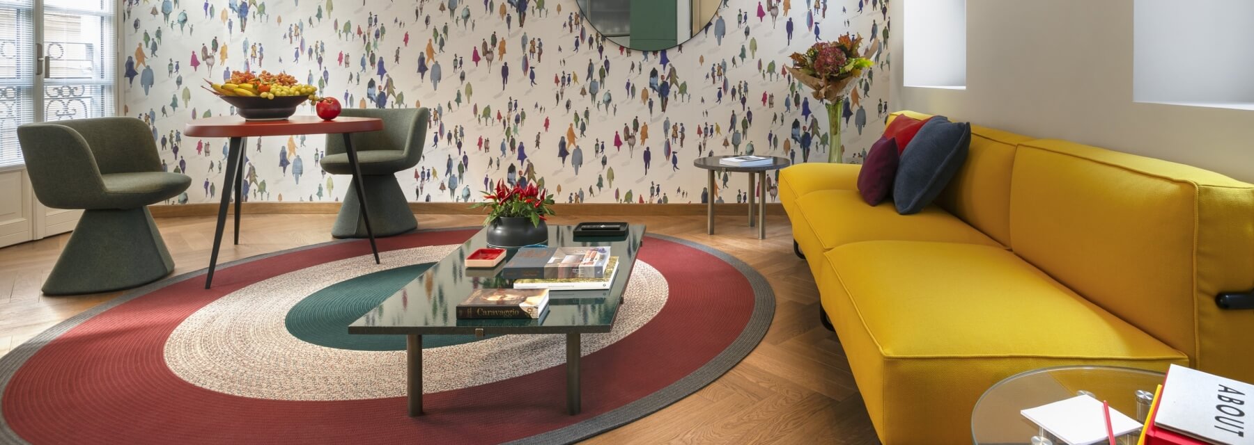 A cozy living room with a yellow couch, circular rug, gray chairs, and eclectic decor, featuring a patterned wall and large mirror.