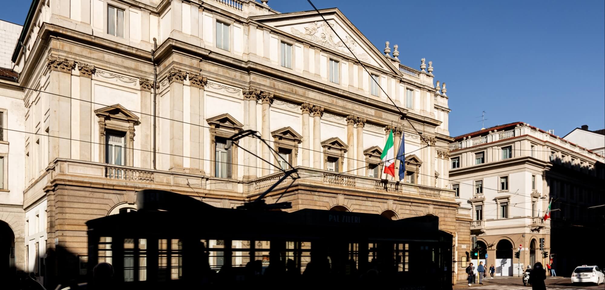 White classical building with Italian flag, people walking, tram tracks, and a clear blue sky.