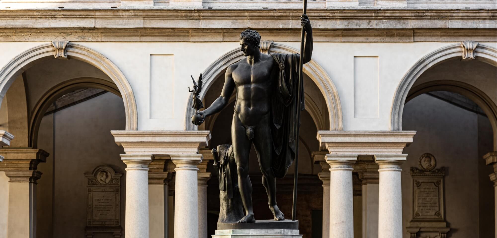 Statua bronzea di un uomo con torcia e bastone, davanti a un edificio bianco ad archi, in un'atmosfera crepuscolare.