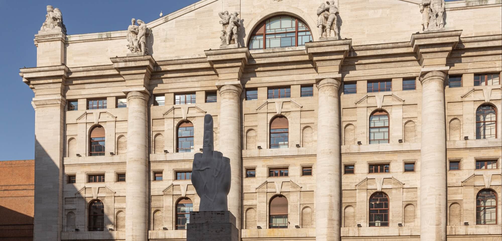 A large classical stone building with columns and a provocative dark stone middle finger sculpture in front, under a blue sky.