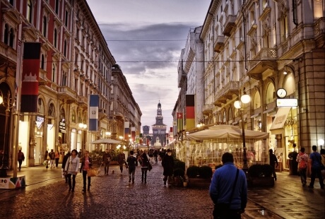 A bustling city street at dusk, filled with people, vibrant buildings, and a prominent clock tower, creating a lively atmosphere.