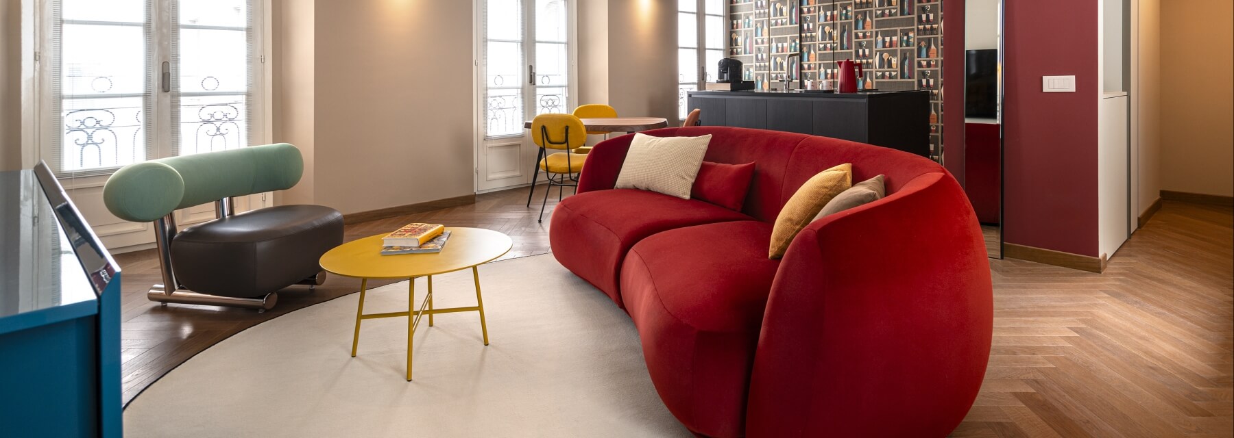 Modern living room with a red couch, yellow coffee table, diverse decor, and natural light from four windows.