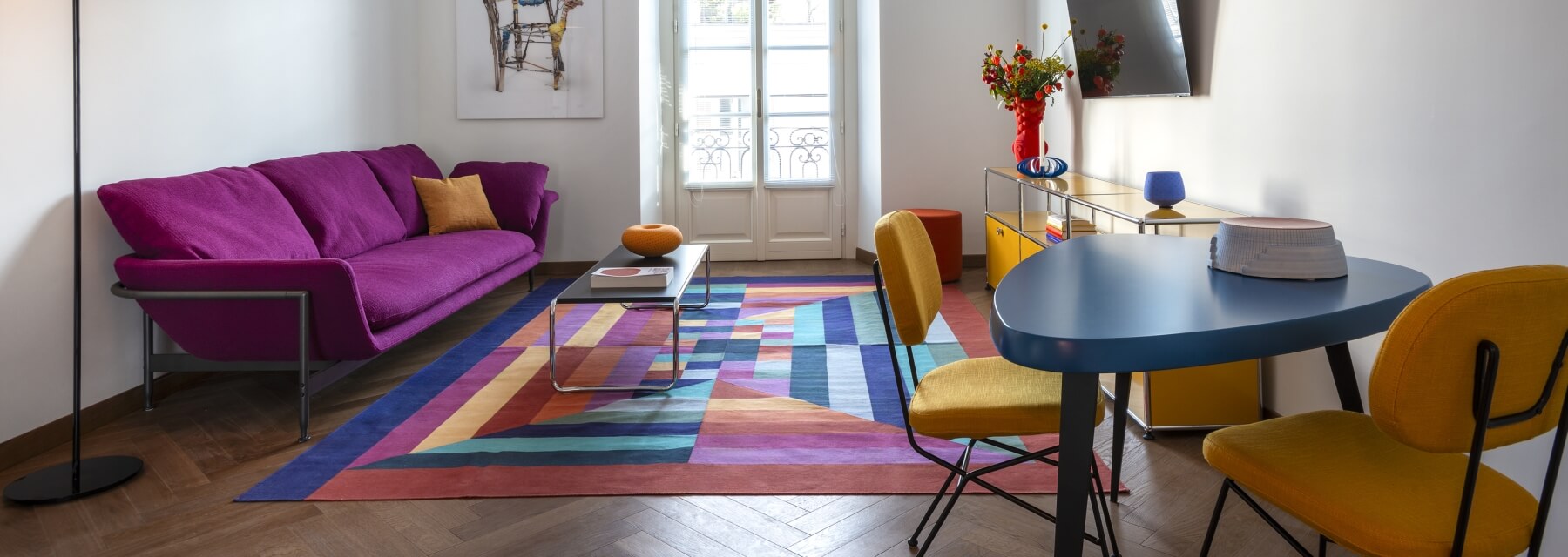 A vibrant living room with a purple couch, blue table, multicolored rug, and natural light from windows.