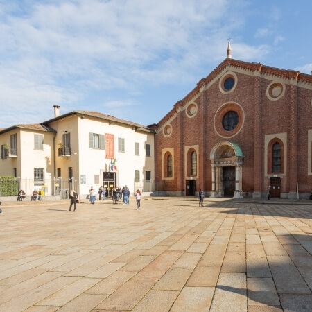 A large red brick building stands in a plaza with smaller structures, under a blue sky with white clouds and scattered people.