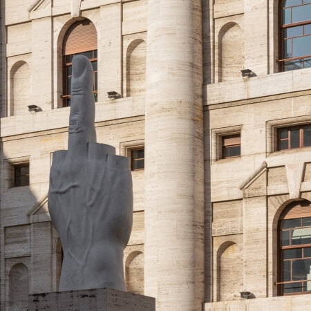 Gray stone sculpture of a middle finger in front of a beige building with windows and columns.