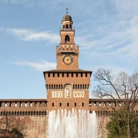 A large stone structure with a clock tower and fountain, surrounded by trees under a blue sky with wispy clouds.