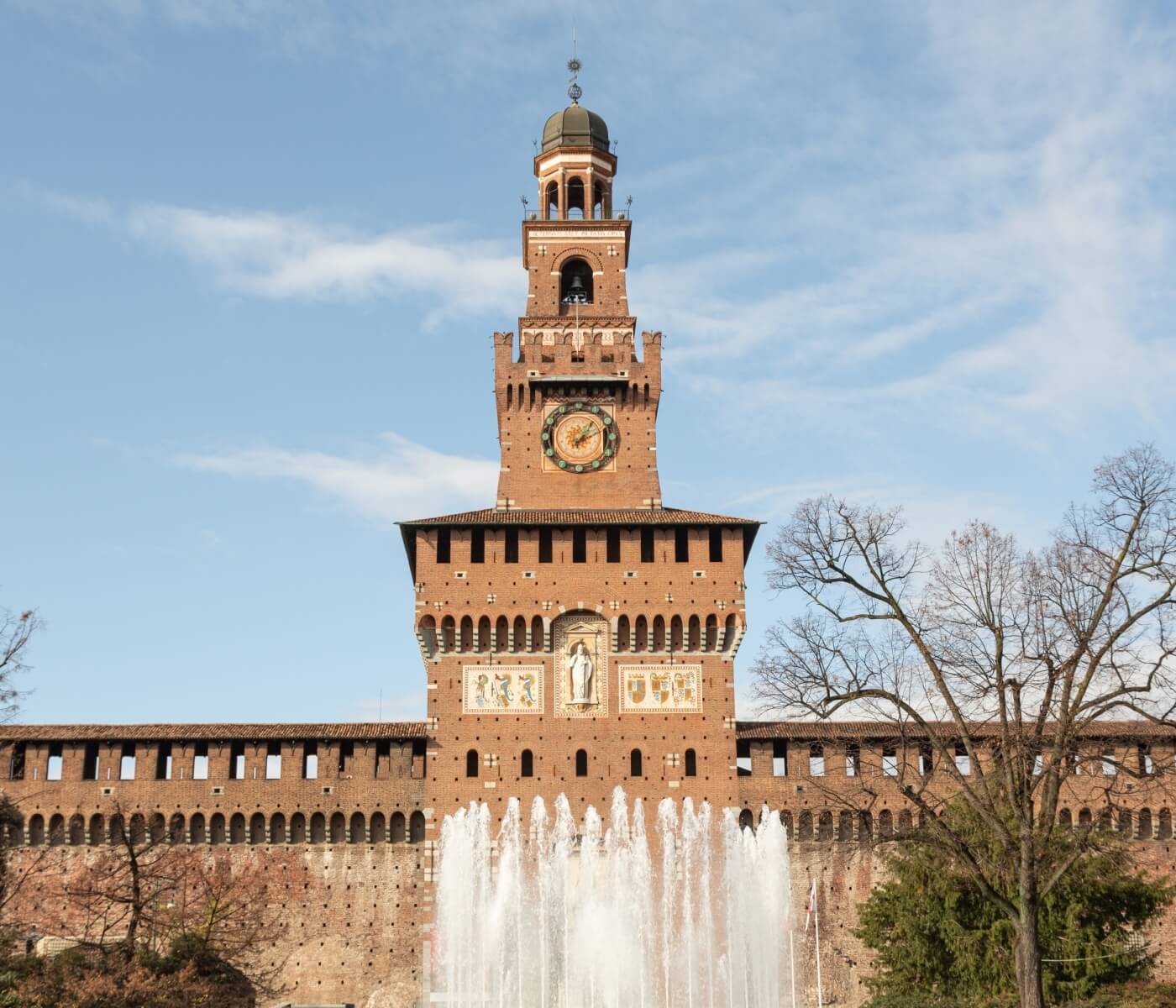 Castello maestoso con torre dell'orologio, fontana e alberi spogli, sotto un cielo sereno e nuvoloso.