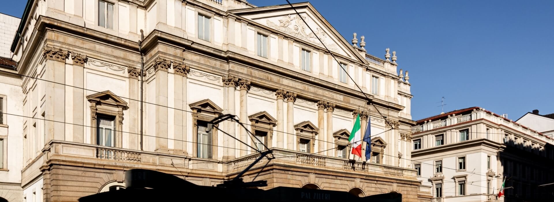 Large white stone building with triangular roof, balcony, flags, and power lines against a clear blue sky.