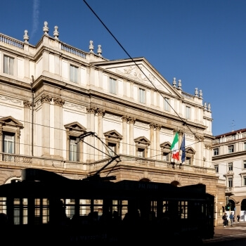 Edificio ornato con tram in silhouette, bandiera italiana, cielo blu e atmosfera vivace di una città europea.