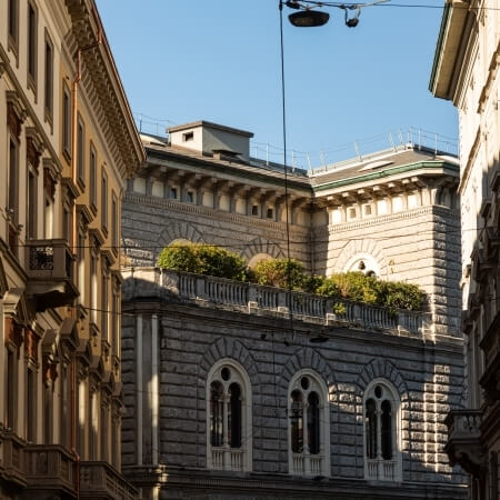 A light-colored stone building with an ornate facade and rooftop greenery stands between two other structures under a blue sky.