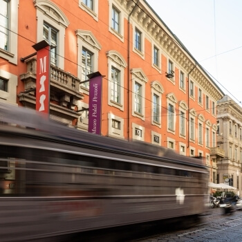 Un tram scuro attraversa una zona storica, con bandiere colorate su un edificio ruggine e un cielo azzurro sopra.