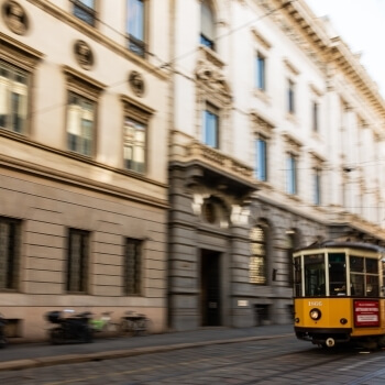 Tram giallo sfocato su strada acciottolata, con edifici beige e bianchi sullo sfondo e oggetti sfocati sul marciapiede.