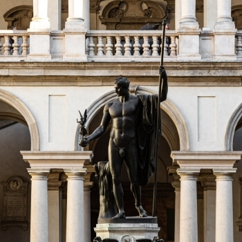 Statua di un uomo con arco davanti a un edificio bianco con colonne e archi, illuminata dalla luce naturale.