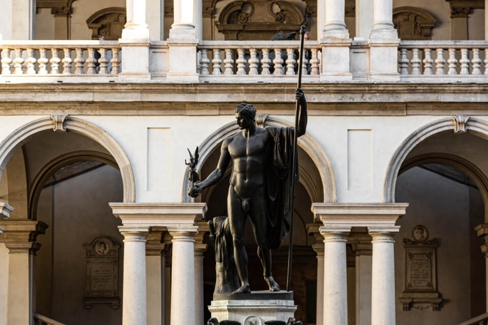 A large white building with archways and a dark metal statue of a nude man holding a spear and a deer's head in front.