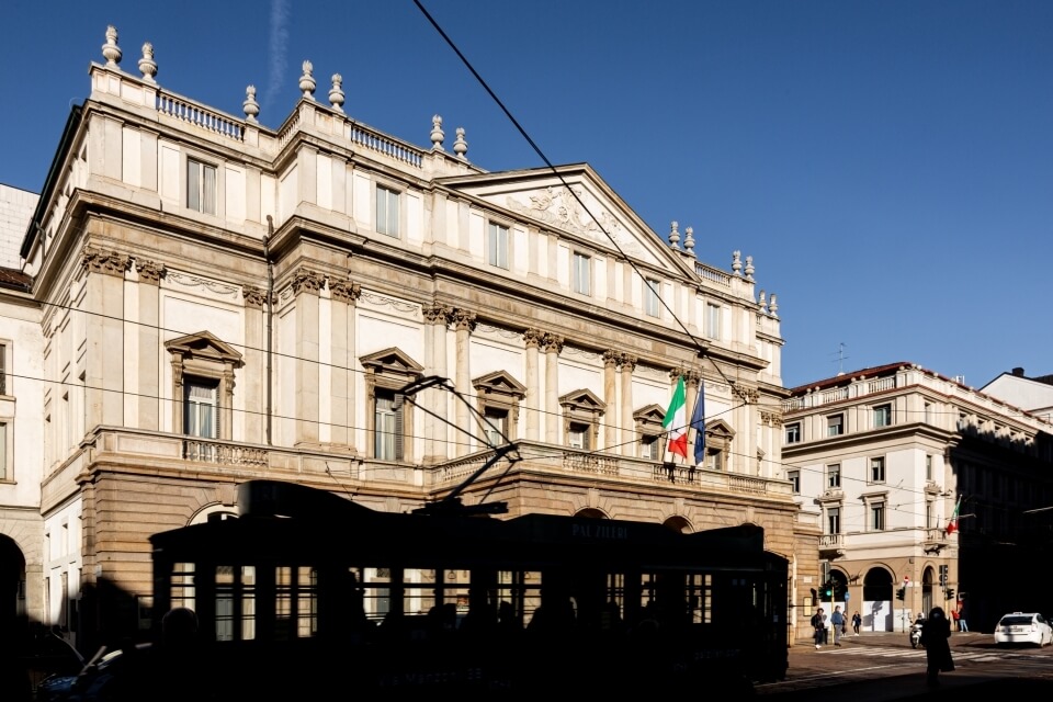 Edificio multi-storico con colonne e bandiere, tram in primo piano, cielo blu e persone che passeggiano.