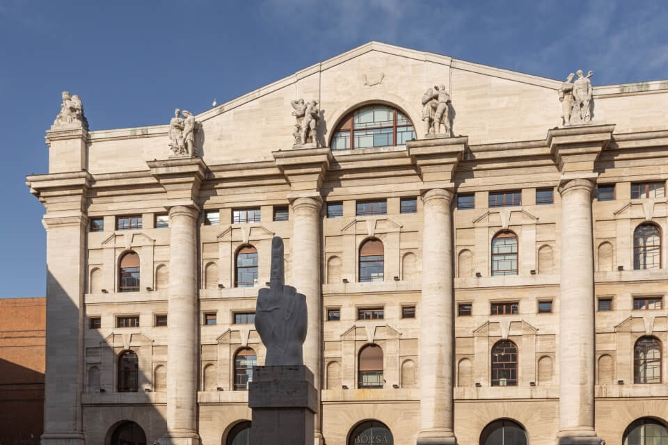 Large stone building with columns and statues, featuring a gray statue of a raised middle finger in front, under a blue sky.