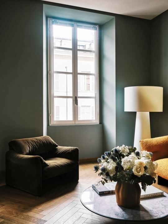 Una comoda sala con un poltrona in pelle marrone, tavolino in vetro, fiori bianchi e un divano arancione sullo sfondo.